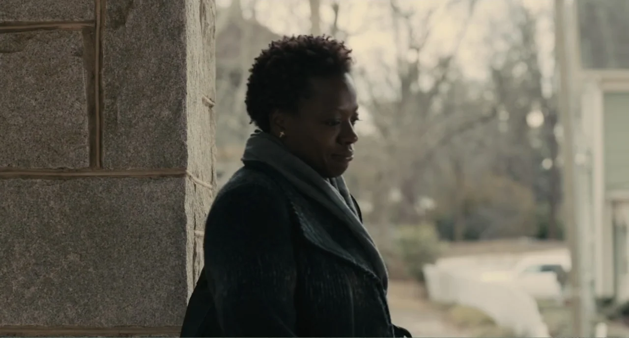Lila & Eve (2015) movie still: medium — A woman stands near a textured granite pillar on an outdoor residential porch area. She is dressed in a dark…