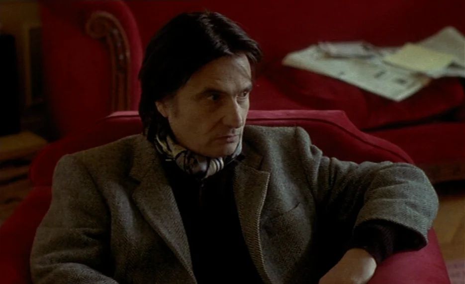 Irma Vep (1996) movie still: medium — A man sits in a vibrant red armchair in a room with hardwood floors. A discarded newspaper rests on a second…