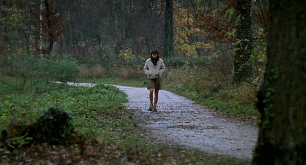 Tiresia (2003) movie still: fullbody — A wet, narrow dirt path winds through a dense autumn forest with fallen leaves. A young woman walks toward…