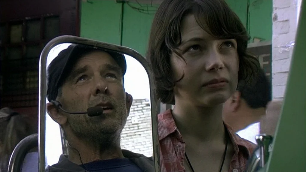 Land of Plenty (2004) movie still: medium — A woman stands near an outdoor stall with a vibrant green wall in background. A man is visible through a…