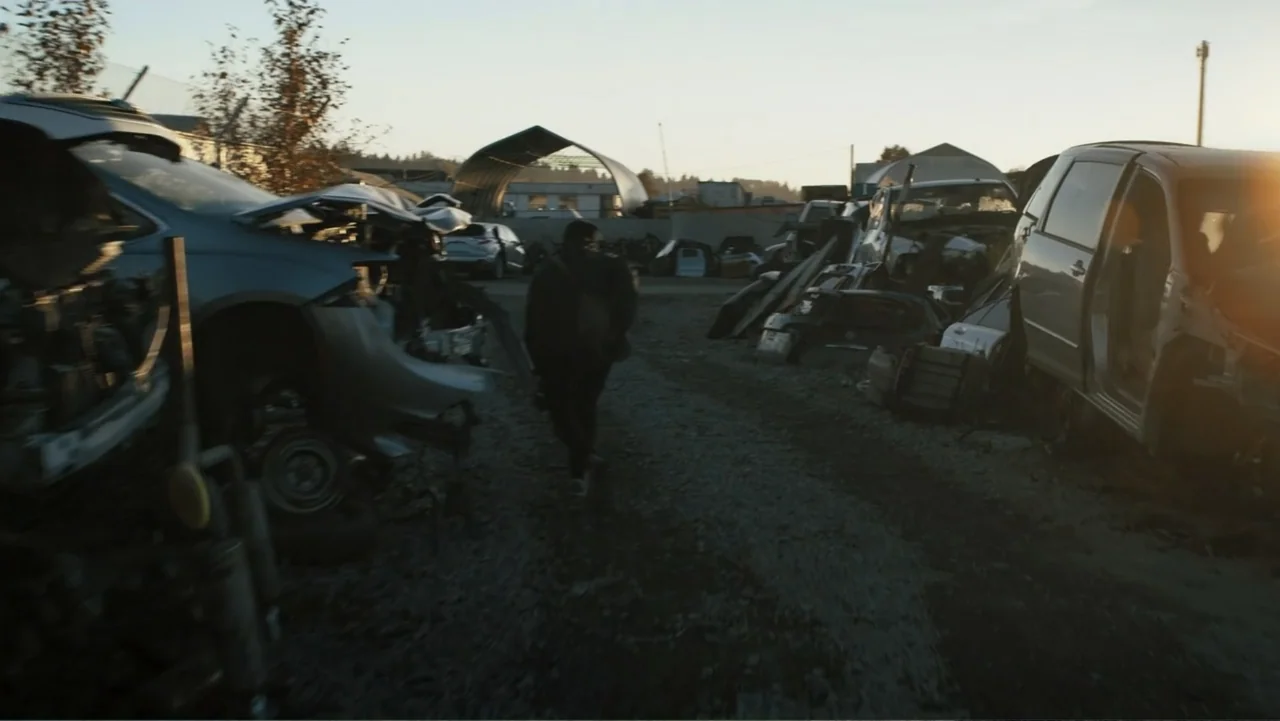 Bullet Proof (2022) movie still: wide — An outdoor car salvage yard contains rows of wrecked vehicles under a pale sky. A lone figure walks along a…