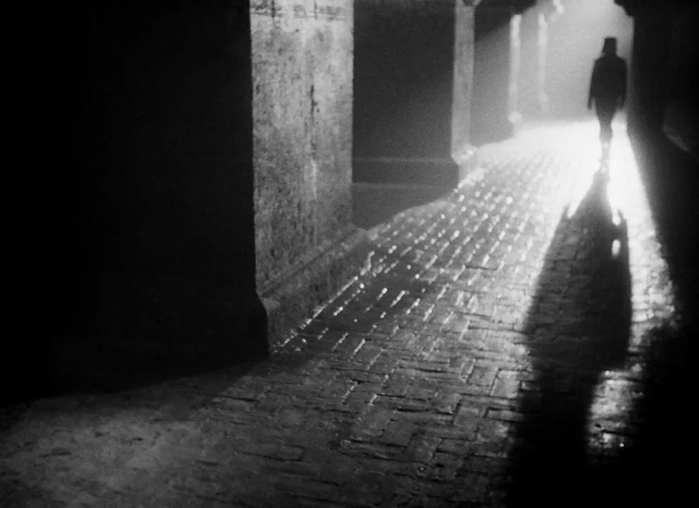 Othello (1952) movie still: wide — A damp underground corridor features large stone pillars and a patterned brick floor. A lone silhouetted person…