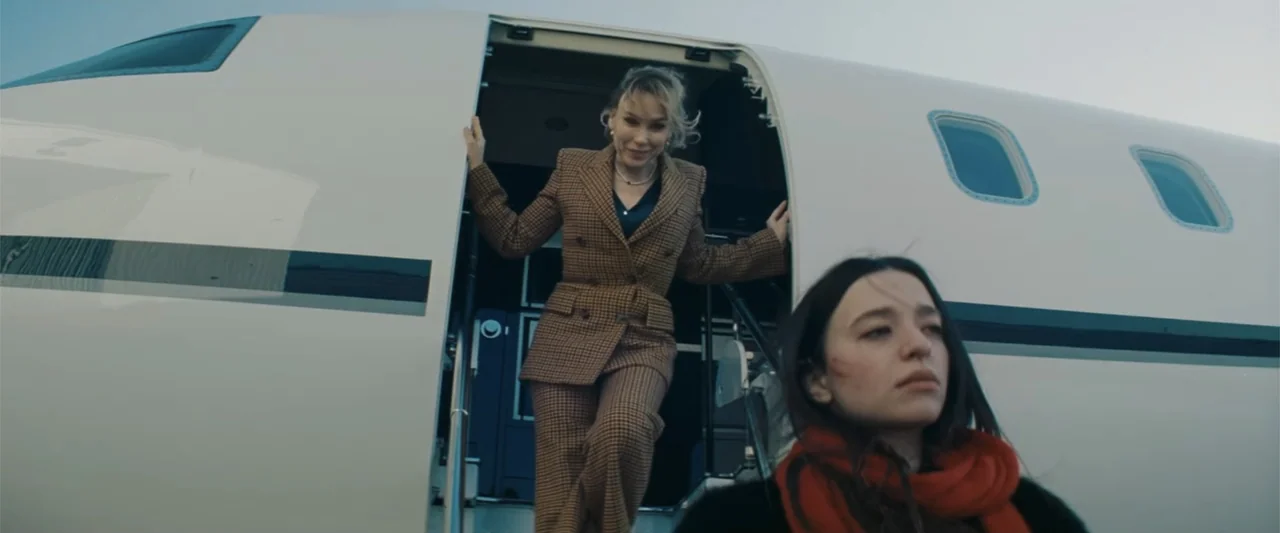 Anora (2024) movie still: fullbody — A private jet sits on the tarmac under a bright, overcast daylight sky. A woman walks down the steps while…