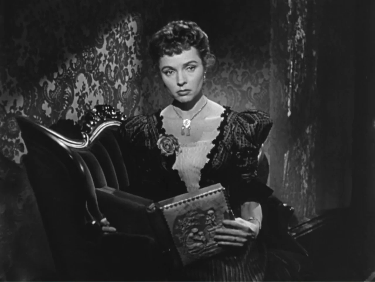 House by the River (1950) movie still: medium — A dimly lit Victorian parlor features ornate wallpaper and a plush, dark upholstered sofa. A woman…