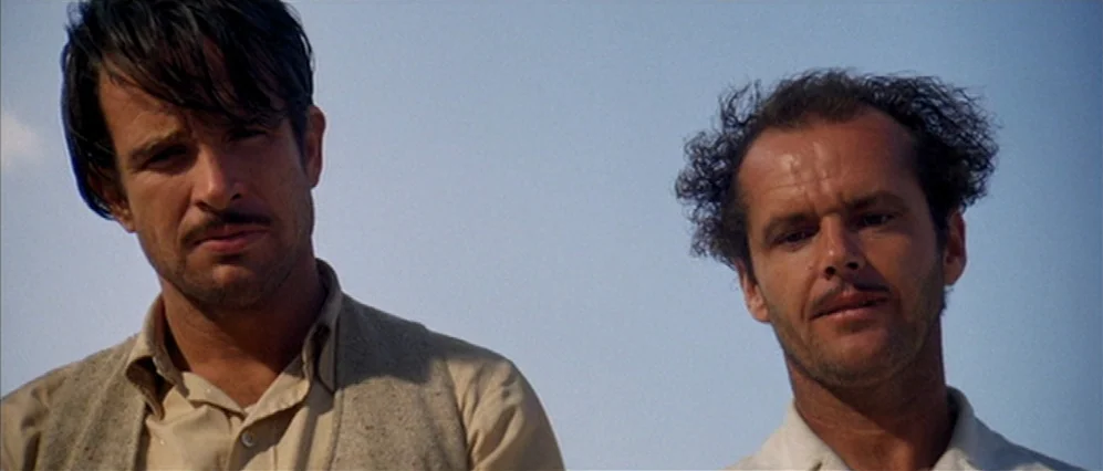 The Fortune (1975) movie still: medium — Two men stand together against a clear blue desert sky background. They are positioned side-by-side looking…