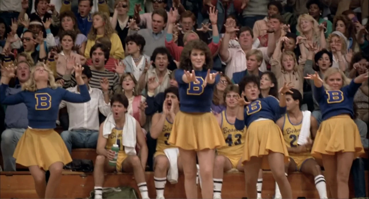 Teen Wolf (1985) movie still: medium — A high school gymnasium with spectators filling wooden bleachers during an athletic event. A group of…