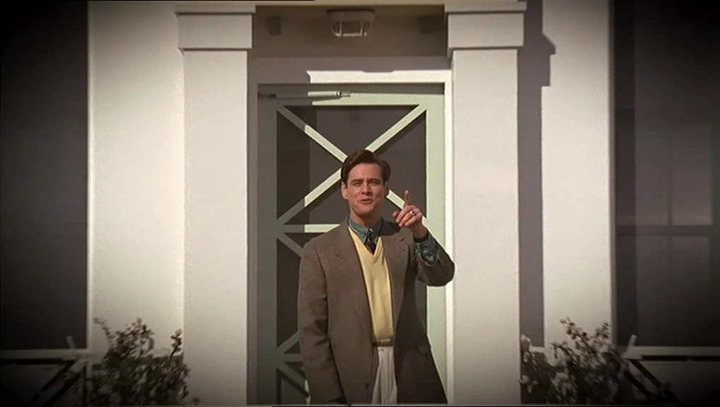 The Truman Show movie still: medium — A man stands in front of a white door with a light green frame. The scene is brightly lit, showing a suburban…