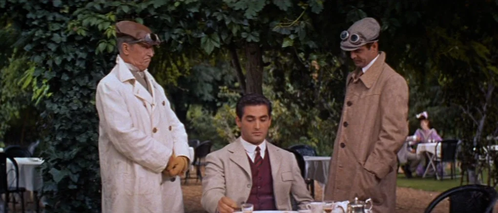 Gigi (1958) movie still: medium — An outdoor restaurant seating area features tables underneath a thick canopy of green leaves. Three men occupy the…