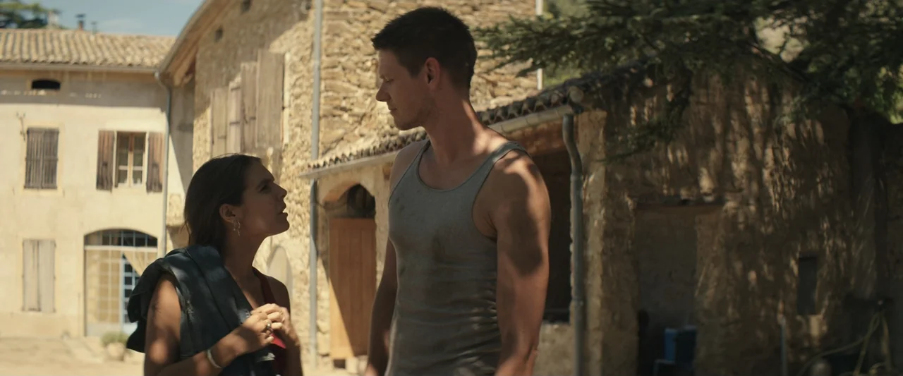 The Only One (2021) movie still: medium — An exterior courtyard with rustic stone buildings and dappled sunlight cast by overhanging trees. A woman…