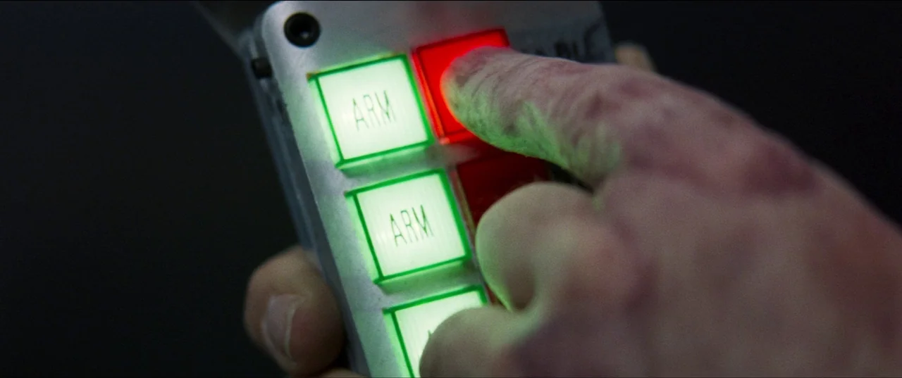 Speed (1994) movie still: closeup — A close view of a hand holding a metallic control device with several buttons. The finger presses a red button…