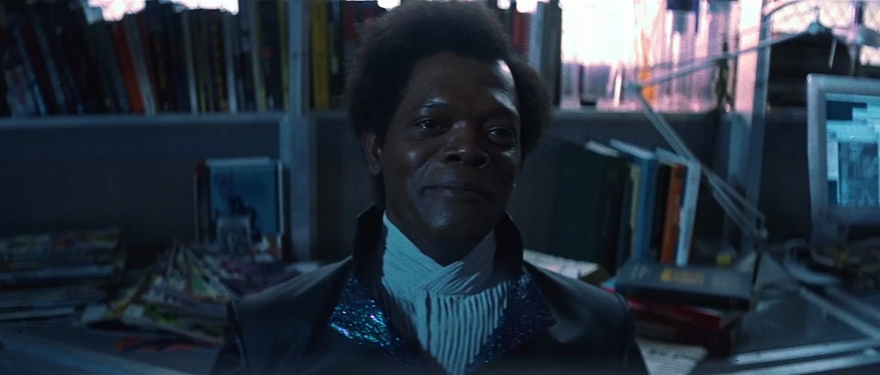 Unbreakable (2000) movie still: medium — A cluttered study contains shelves packed with books and a computer monitor glowing blue. A man with an…
