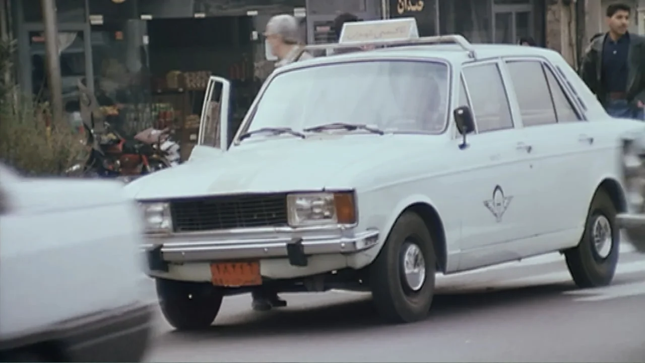 The Mirror (2014) movie still: medium — A white sedan functions as a taxi on a paved city street during daytime. The car moves forward past…