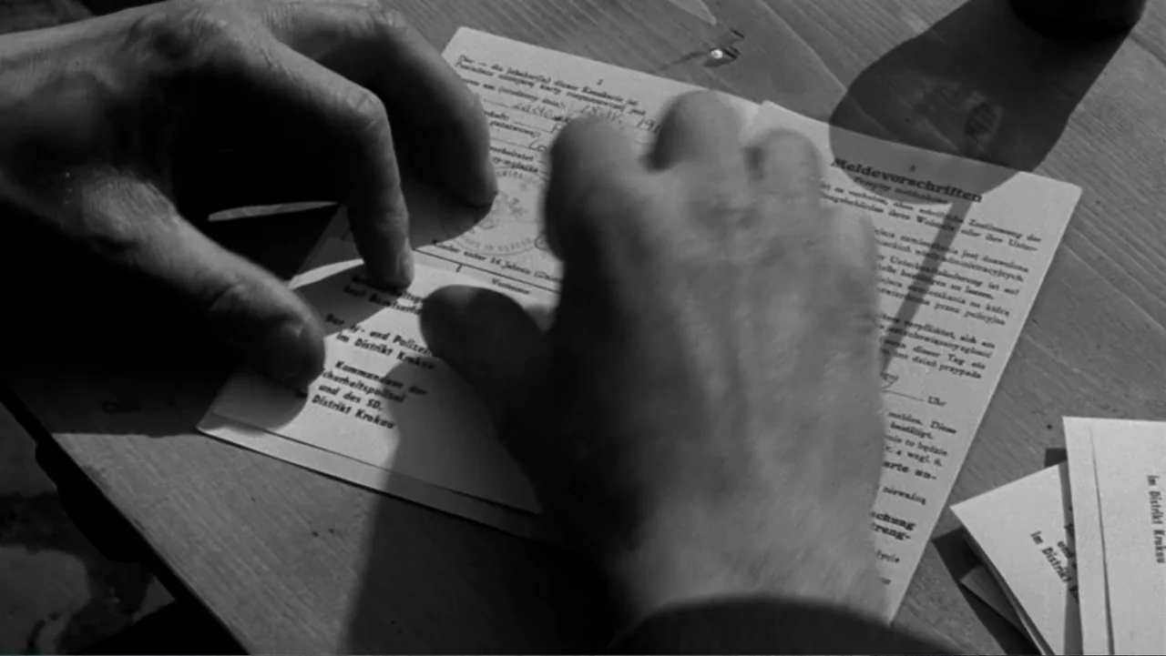Schindler's List (1993) movie still: closeup — A wooden desktop surface sits under bright directional light. Two hands place a printed German…