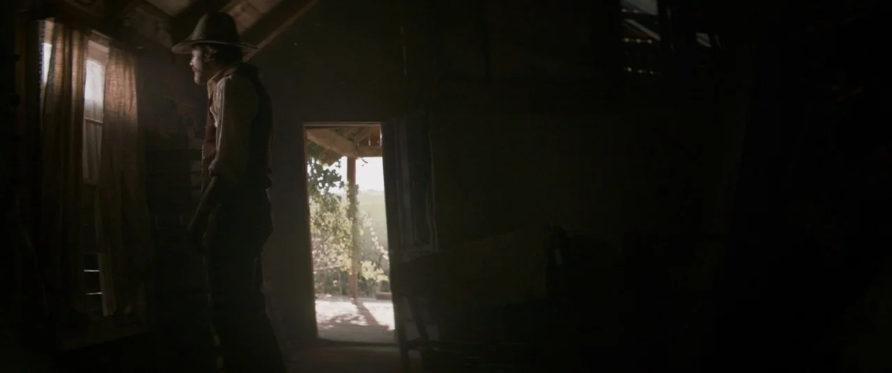 The Sisters Brothers (2018) movie still: medium — A dark wooden room illuminated by daylight filtering through a window and an open door. A man in a…
