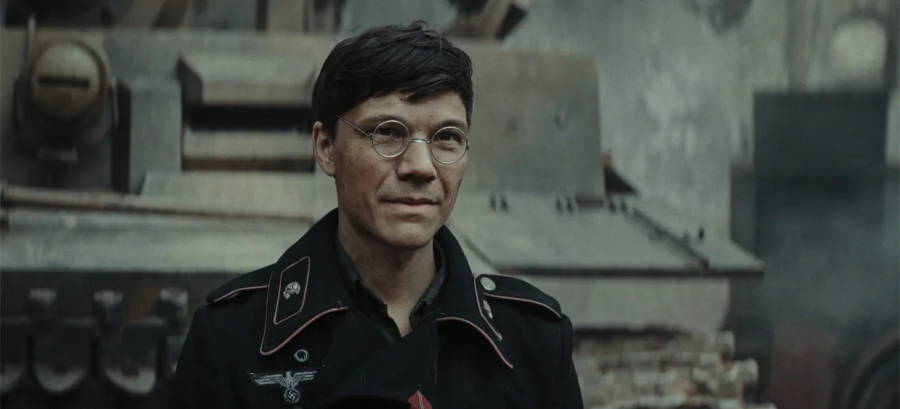 The Tank (2025) movie still: medium — A man in a 1940s military uniform stands in front of a heavy industrial vehicle. He faces the camera while…