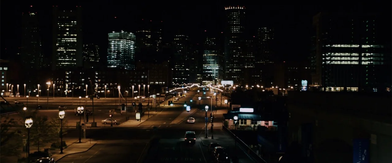 Zookeeper (2011) movie still: establishing — A wide view of a deserted city intersection at night featuring skyscrapers. A few cars drive along the…