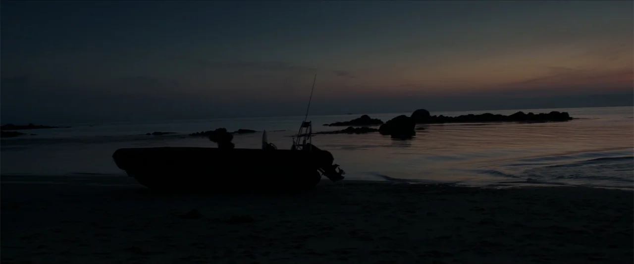 Apocalypse Z: The Beginning of the End (2024) movie still: wide — A dark, calm shoreline with rocks in the water and a boat parked nearby. The…