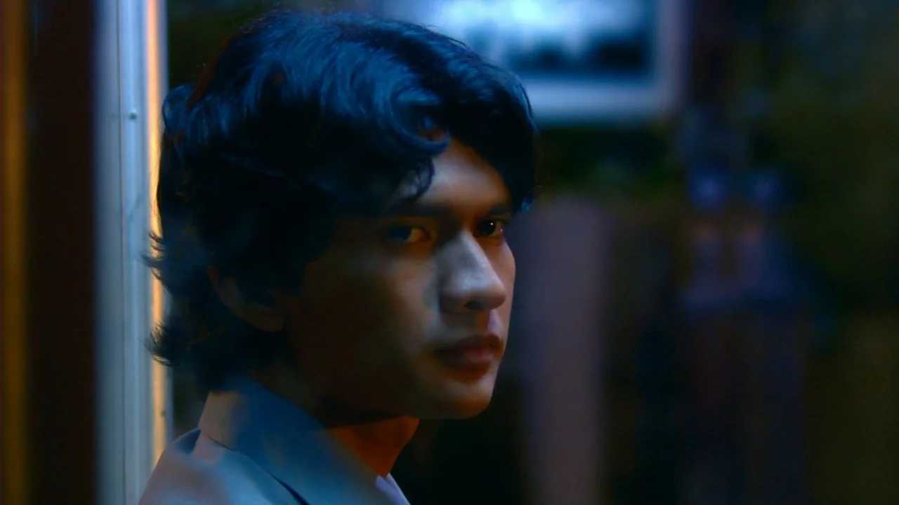 Merantau (2009) movie still: closeup — A man in a shirt leans against a metal pillar inside a dimly lit train car. He gazes forward while blue neon…