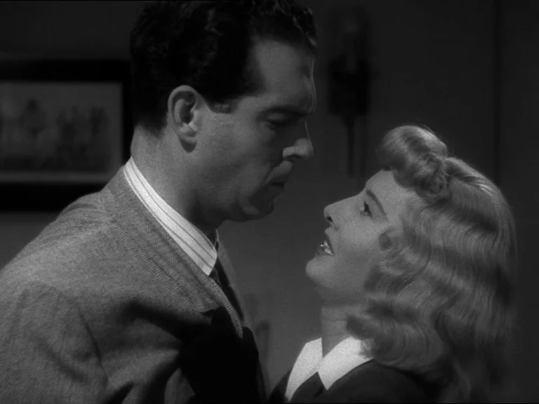 Double Indemnity (1944) movie still: medium — A dark indoor setting with a blurred framed picture hanging on a wall. A man and a woman stand face to…
