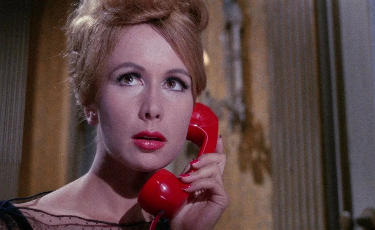 Blood And Black Lace (1964) movie still: closeup — A dimly visible interior space features textured golden-yellow wallpaper behind the subject. A…