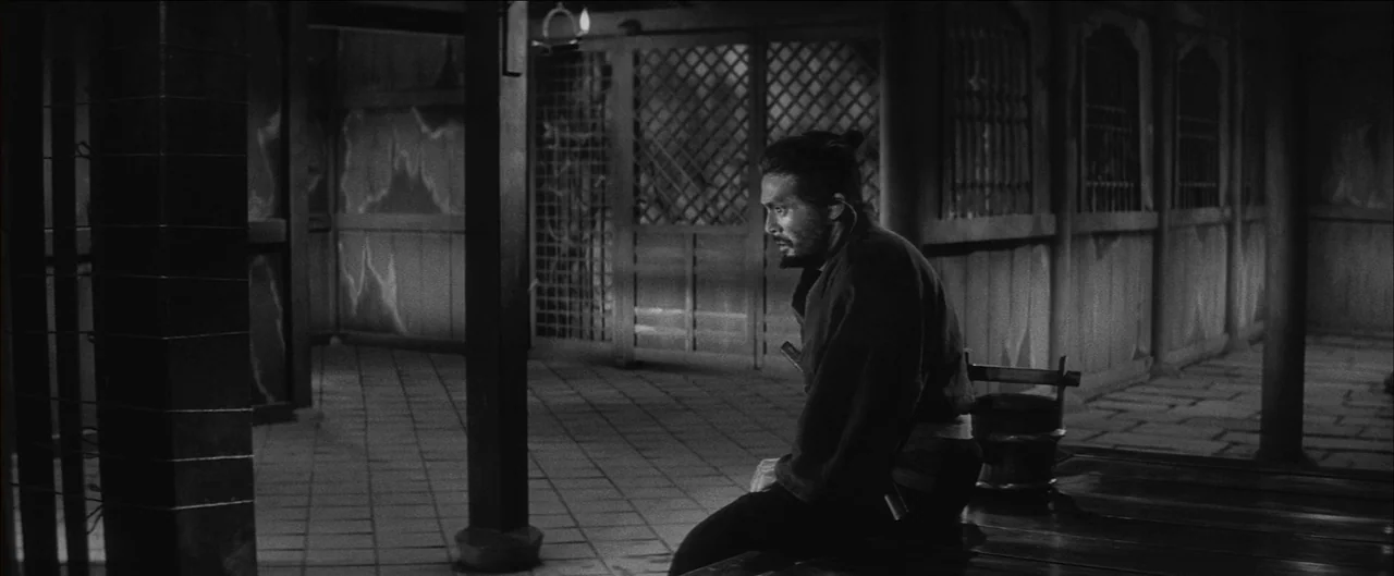 Harakiri (1962) movie still: medium — A dimly lit wooden structure with latticed panels and a stone-tiled floor at night. A lone man with a topknot…