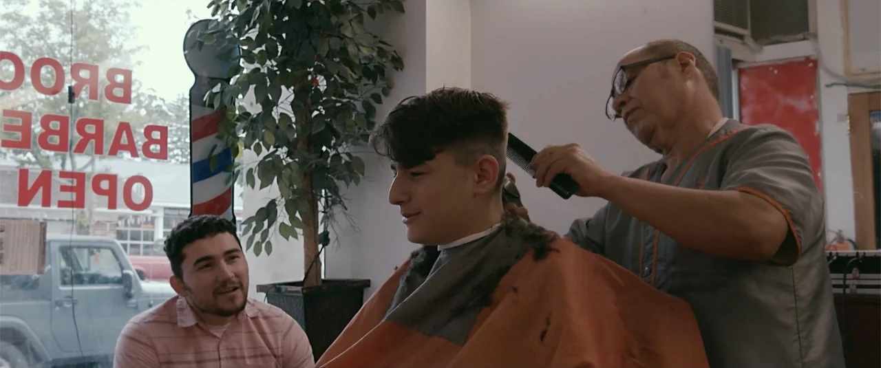 Simple as Water (2021) movie still: medium — A well-lit barbershop interior features a large window with red vinyl signage. A barber grooms a seated…
