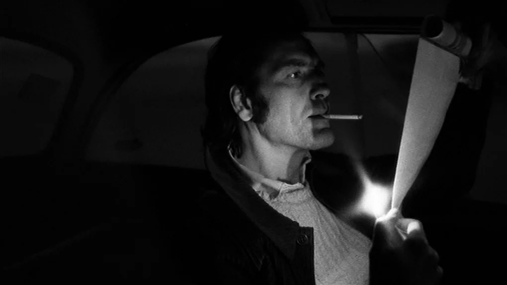 The State of Things (1982) movie still: medium — The interior of a dark car at night. A man reads a document illuminated by a cigarette lighter flame.