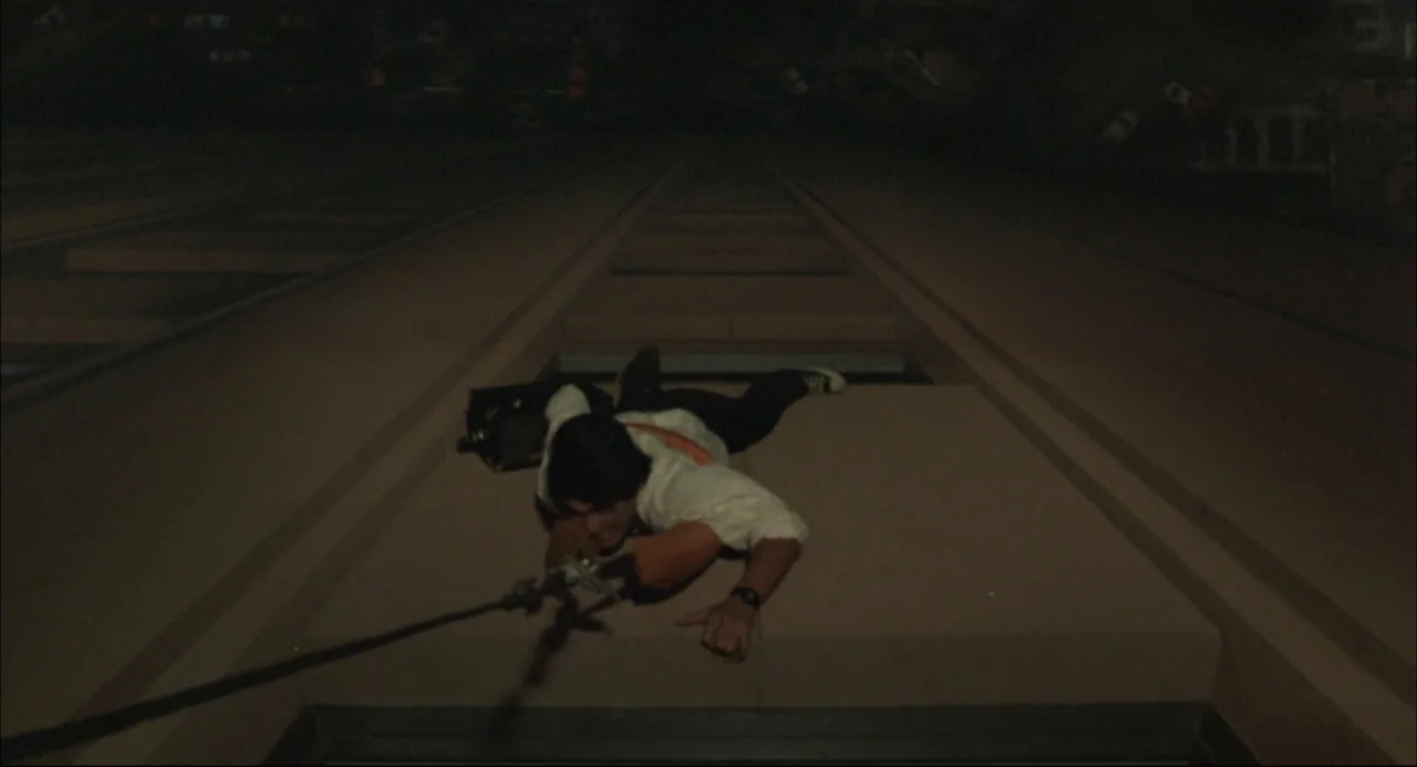 The Return of Godzilla (1984) movie still: wide — The flat top of a moving train carriage stretches into the dark night. A young man lies prone,…