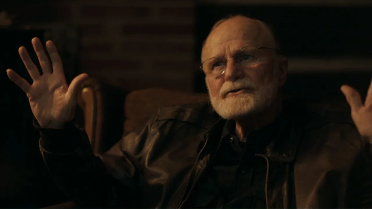 Alabama Snake (2020) movie still: medium — A dark, dimly lit interior room features a brick wall in the background. An older man with a white beard…