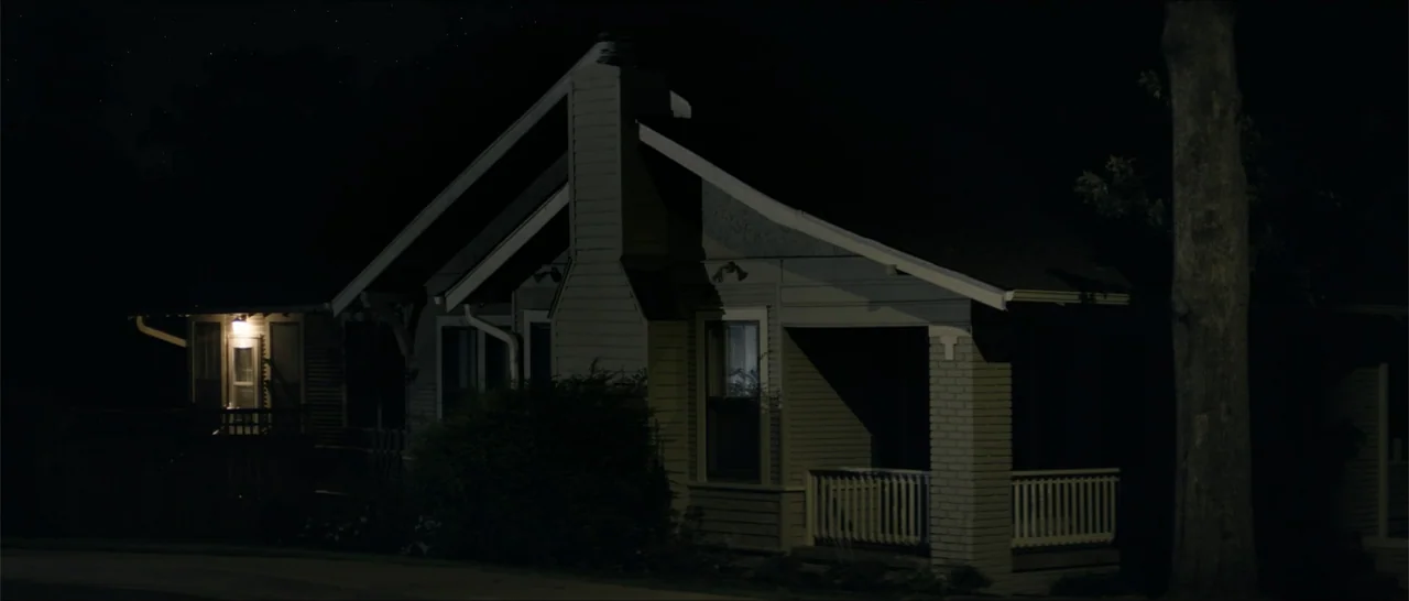 Crescent City (2024) movie still: establishing — A one-story residential house is silhouetted against a dark sky at night. The structure is shrouded…