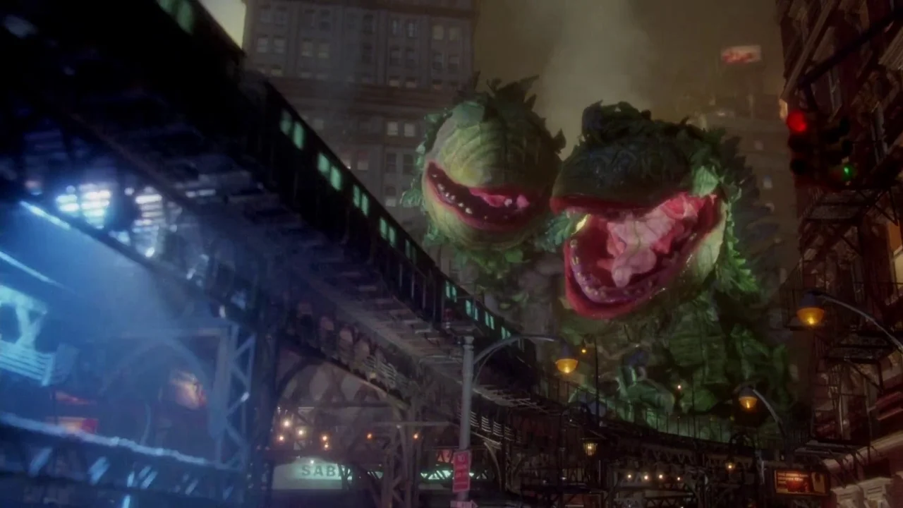 Little Shop of Horrors (1986) movie still: wide — An urban cityscape at night features a towering elevated train track structure. Two massive,…