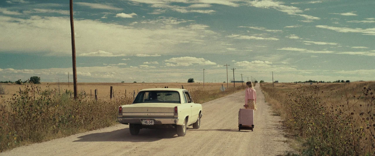 LaRoy, Texas (2023) movie still: wide — A flat, rural landscape stretches toward a distant horizon under a cloudy blue sky. A young woman walks…
