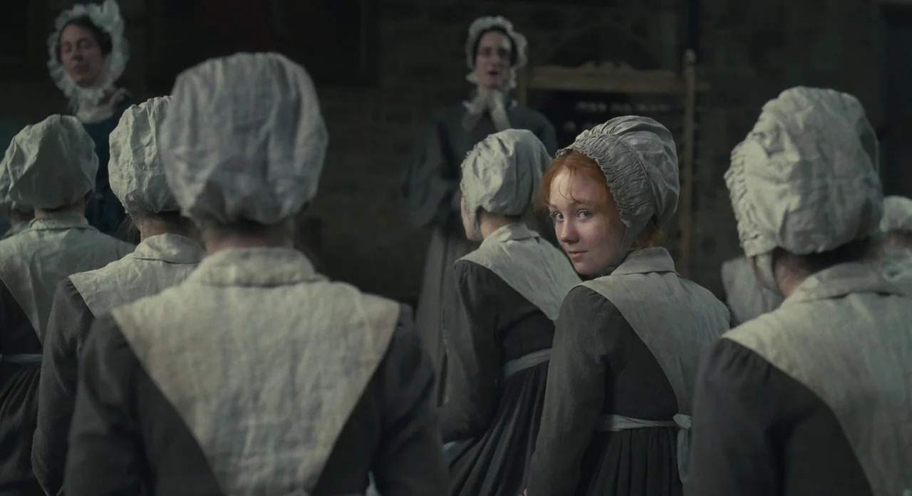 Jane Eyre (2011) movie still: medium — A group of women in 19th-century modest attire sit in a dim stone room. One girl turns around to look…