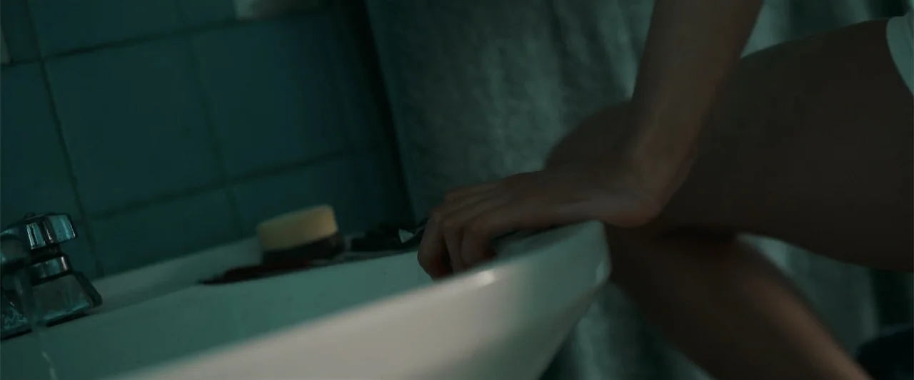 Evil Eye (2020) movie still: closeup — A dimly lit bathroom features a porcelain sink with a bar of soap. A person rests their hand on the sink edge…