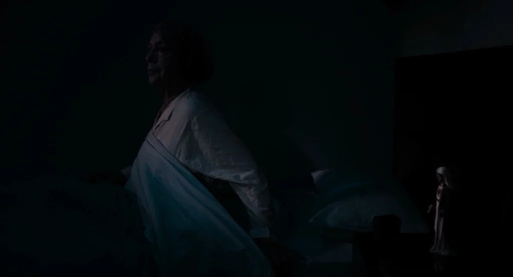 Lourdes movie still: medium — A dark bedroom interior with a woman sitting upright in a bed. A small religious figurine stands on the nightstand…