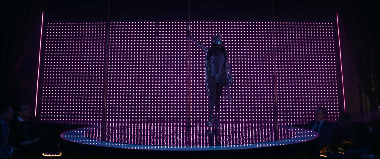 Hustlers (2019) movie still: wide — A dimly lit club interior features a central circular stage with a pole. A woman in a sequined costume stands…