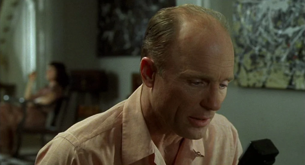Pollock (2000) movie still: medium — A man sits in the foreground with a blurred woman seated in the background. Abstract artwork hangs on the walls…