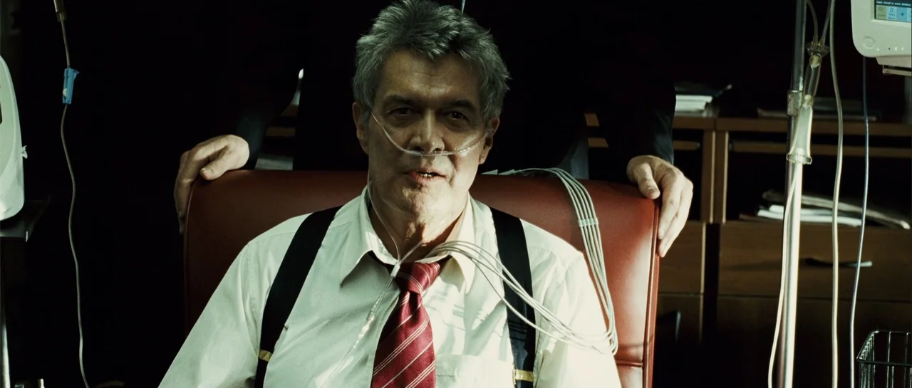 Shoot 'Em Up (2007) movie still: medium — A man sits in a leather chair within a professional medical office setting. The man wears a nasal cannula…