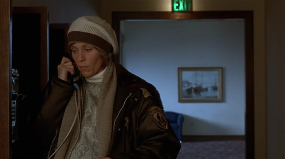 Fargo (1996) movie still: medium — A dimly lit indoor space featuring a beige wall, a framed painting, and an illuminated exit sign. A woman stands…