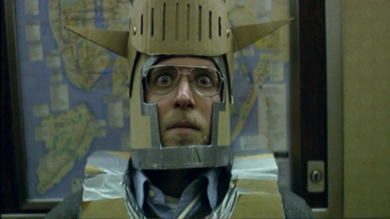 Murder Party movie still: closeup — A man stands inside a brightly lit, small elevator space. He wears a makeshift suit of cardboard and foil with a…