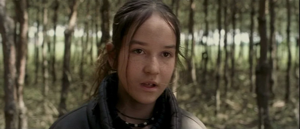 Time Of The Wolf (2003) movie still: closeup — A dense grove of pine trees with thin, vertical trunks fills the background. A young girl with her…