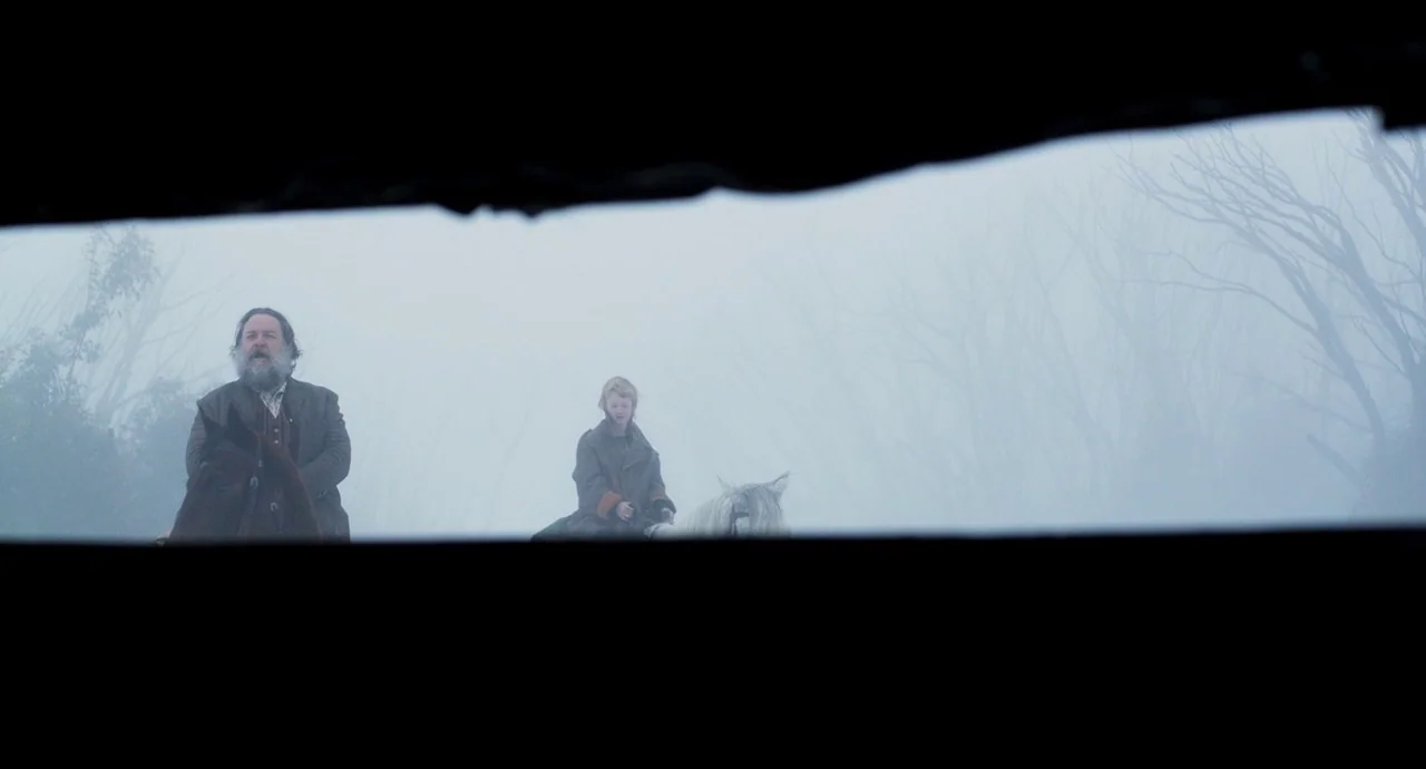 True History of the Kelly Gang (2019) movie still: wide — Two men ride horses through a dense, grey fog in an open outdoor expanse. Bare trees are…
