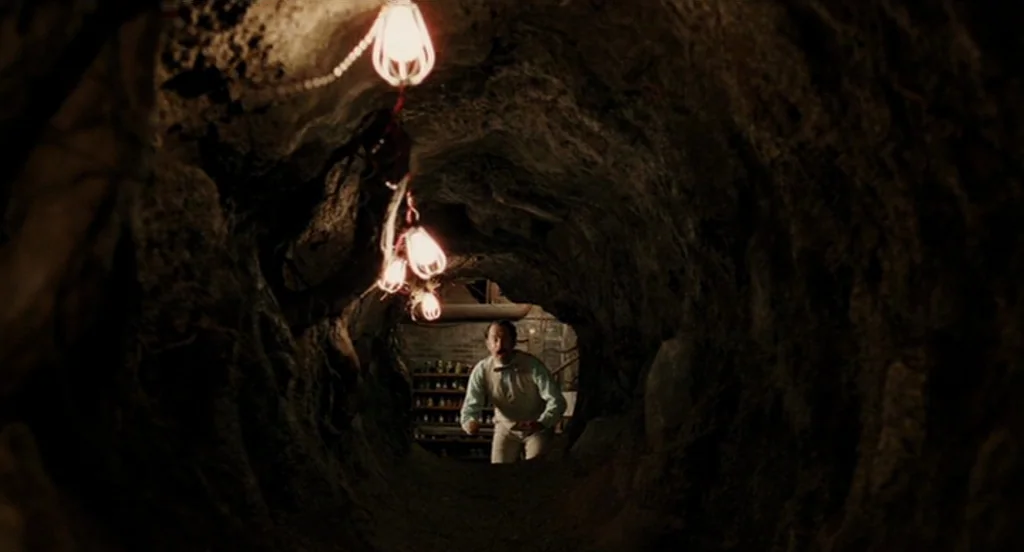 The Ladykillers (2004) movie still: wide — A dark, cramped, dirt-walled tunnel lit by a series of dangling wire-cage lamps. A man in a dress shirt…