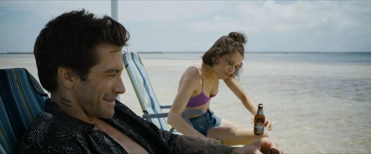 Road House (2024) movie still: medium — Two people sit in beach chairs on a shallow sandy shore under a bright sky. The man watches the woman as she…