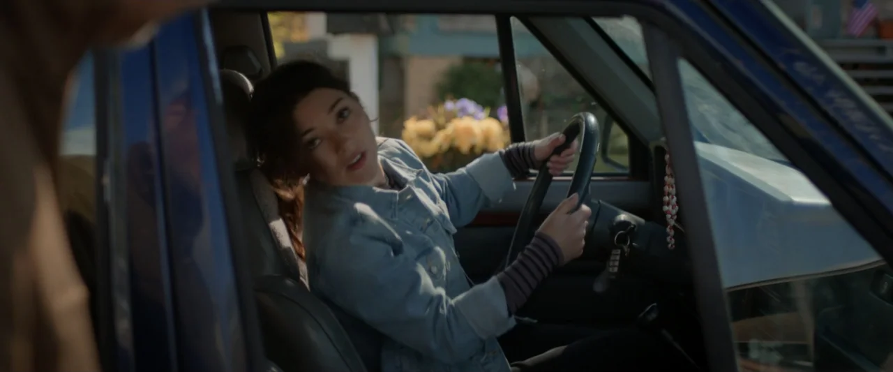 Clown in a Cornfield (2025) movie still: medium — A woman sits in the driver seat of a car looking out the window. An obscured figure stands outside…