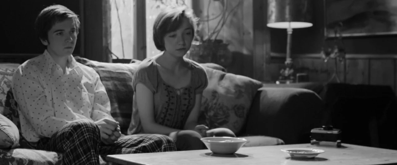 Weirdos (2016) movie still: medium — A living room interior with wood-paneled walls and a fabric-covered sofa. Two young adults sit together on the…