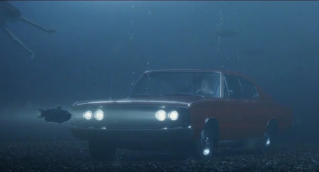 Big Fish (2003) movie still: wide — An underwater scene shows a vintage red sedan resting on a dark, silty bed. Headlights illuminate the murky…