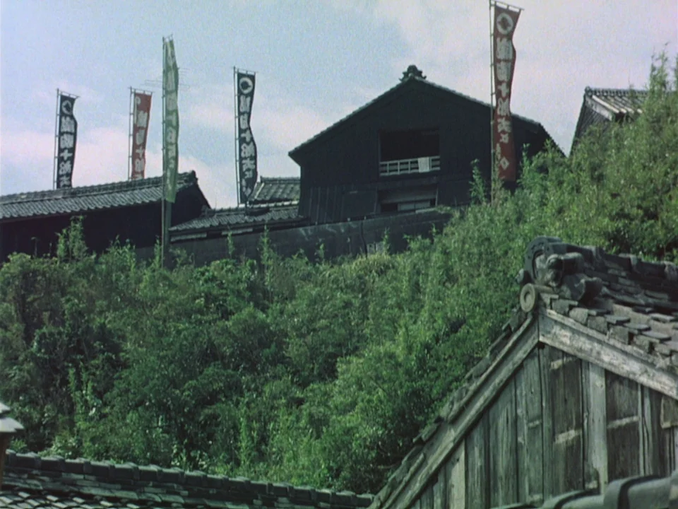 Floating Weeds (1959) movie still: establishing — Traditional dark wooden Japanese buildings sit atop a hill covered in dense green foliage. Tall…