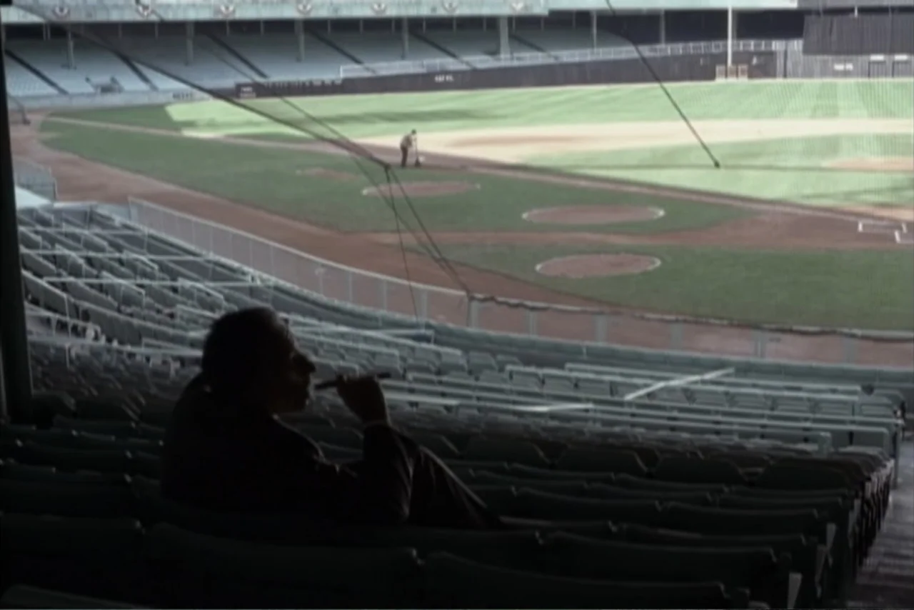 Unification of Japan 61 (2024) movie still: wide — An expansive empty sports field viewed from the upper deck of a grandstand. A man sits alone in…