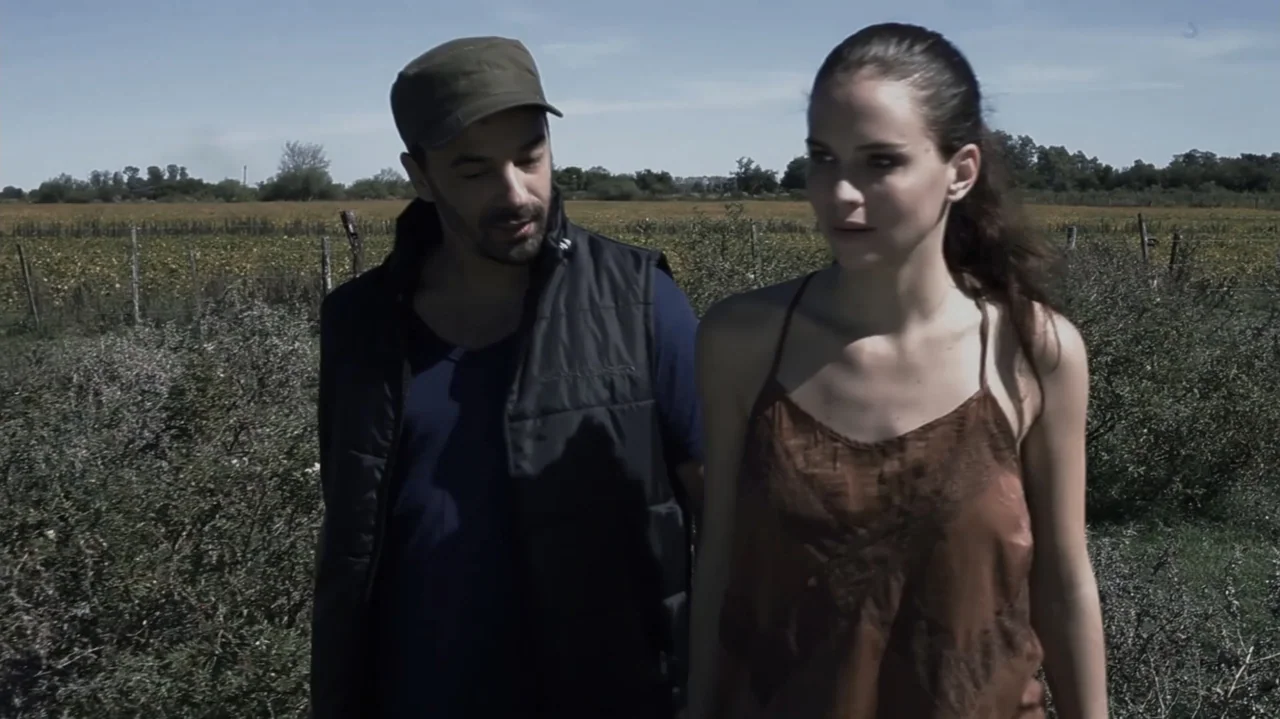 Still Life (2013) movie still: medium — An open rural field with sparse dry vegetation under a clear, bright daylight sky. A man in a cap and a…